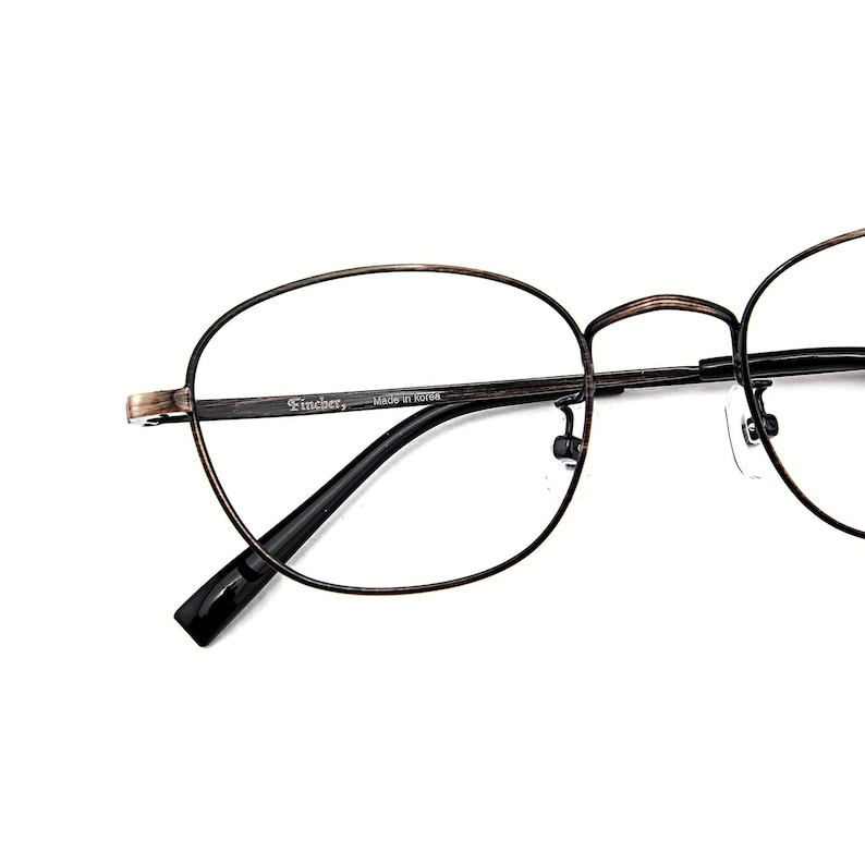 May include: Oval spectacles with a dark brown metal frame. The glasses have a thin frame and black temples. The brand name "Fincher" is printed on the frame. The lenses are clear.