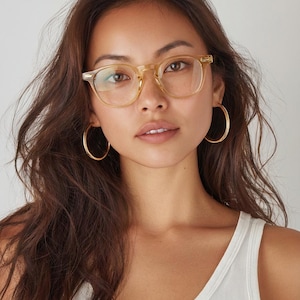 Puede incluir: Una mujer con gafas de montura transparente y pendientes de aro dorados. Las gafas tienen forma rectangular. La mujer lleva una camiseta sin mangas blanca. Su cabello casta&ntilde;o oscuro es largo y ondulado.