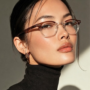 May include: A woman wearing eyeglasses with a brown upper frame and clear lower frame. The woman is wearing a black turtleneck sweater and gold hoop earrings. The background is a neutral color.