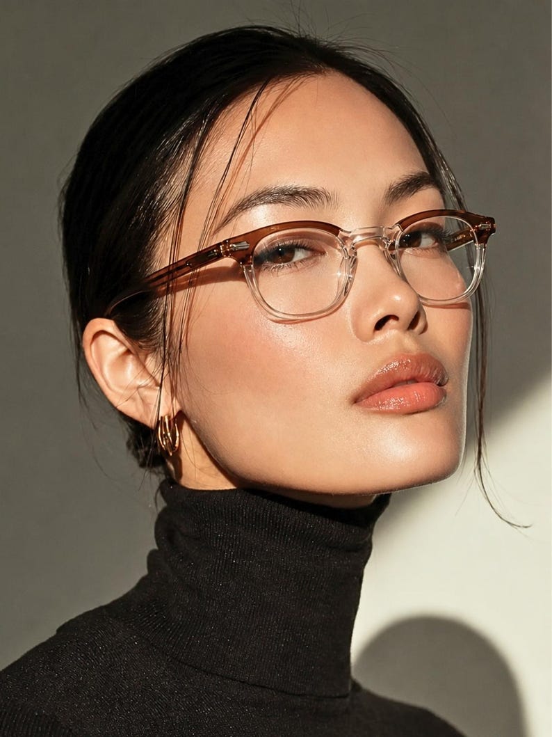 May include: A woman wearing eyeglasses with a brown upper frame and clear lower frame. The woman is wearing a black turtleneck sweater and gold hoop earrings. The background is a neutral color.