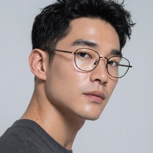 May include: Close-up of a person wearing round, thin-framed spectacles with a bronze finish. The spectacles have a classic design. The person is wearing a dark grey t-shirt.