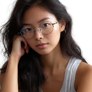 May include: Round-framed eyeglasses with thin, dark metal rims and clear lenses. The frames feature a minimalist bridge and slender temples, providing a classic, lightweight look. These glasses are designed with a simple, circular shape that rests comfortably on the face, offering a clear view through the transparent lenses.