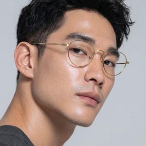 May include: Close-up of gold-rimmed eyeglasses with a round frame. The glasses have a thin, delicate design and are set against a neutral background. The temples have a green accent.
