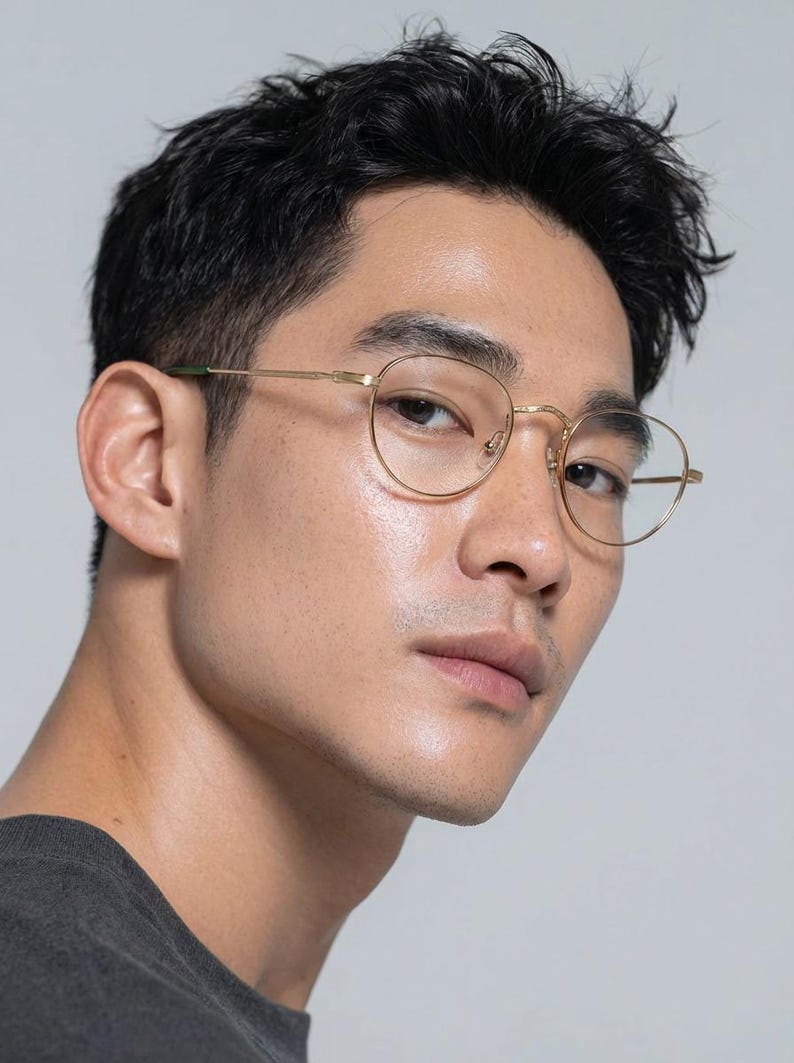 May include: Close-up of gold-rimmed eyeglasses with a round frame. The glasses have a thin, delicate design and are set against a neutral background. The temples have a green accent.
