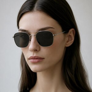 May include: Sunglasses with a thin silver metal frame and a double bridge design. The lenses are dark gray and have a rounded square shape. The temples are slender and metallic, connecting to the front frame with small hinges.