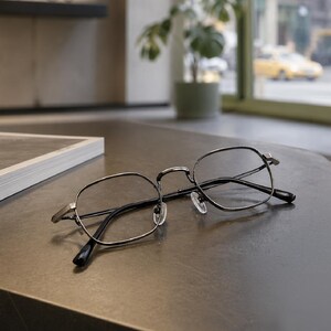 May include: A pair of eyeglasses with thin, dark metallic frames in a rounded hexagonal shape. The glasses feature clear lenses, a slim bridge, and adjustable nose pads, with black temple tips extending from the metallic arms of the frame.