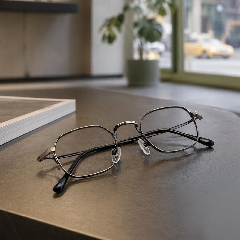 May include: A pair of eyeglasses with thin, dark metallic frames in a rounded hexagonal shape. The glasses feature clear lenses, a slim bridge, and adjustable nose pads, with black temple tips extending from the metallic arms of the frame.