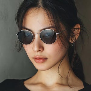 May include: Silver-framed, round sunglasses with dark lenses. The glasses are worn by a person with dark hair and a black top. The background is a solid grey colour.