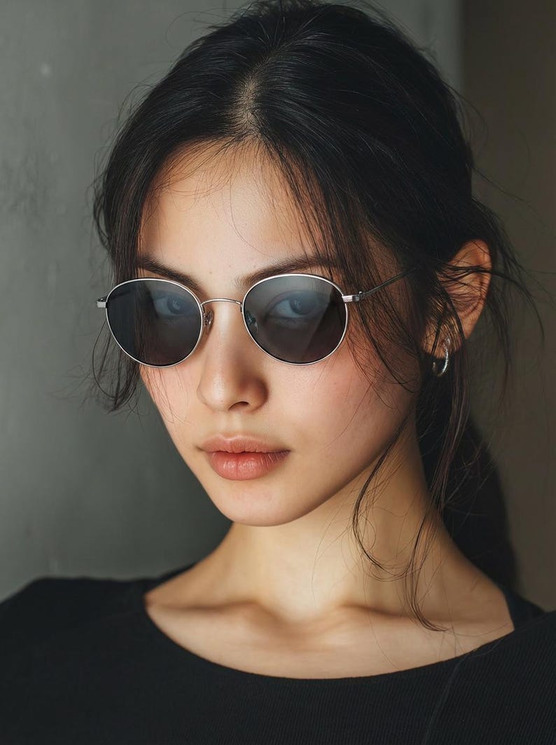 May include: Silver-framed, round sunglasses with dark lenses. The glasses are worn by a person with dark hair and a black top. The background is a solid grey colour.