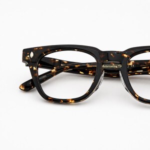 Puede incluir: Primer plano de gafas con un patr&oacute;n carey marr&oacute;n oscuro. Las monturas tienen forma cuadrada con bordes redondeados. El texto "Reinventing Vir" es visible en el puente. Las patillas son de color marr&oacute;n oscuro liso.