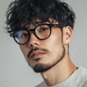 May include: Round black eyeglasses with a dark frame. The glasses are worn by a person with dark curly hair and a beard. The person is wearing a gray crew neck shirt.
