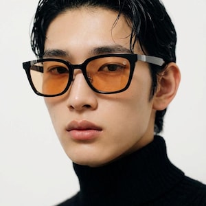 May include: Black square-framed sunglasses with orange-tinted lenses. The glasses have silver accents on the temples and a small logo on the frame. The person is wearing a black turtleneck.