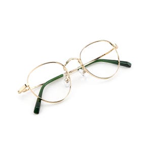 May include: Gold-rimmed eyeglasses with a round frame and green temples. The glasses have a delicate, minimalist design, with a thin gold frame and clear lenses. The temples are a dark green color.