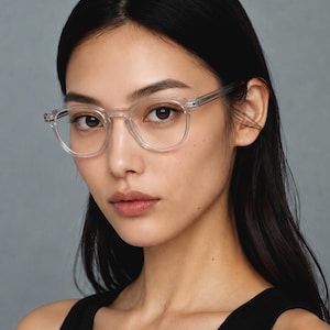 K&ouml;nnte beinhalten: Eine Person tr&auml;gt eine transparente Brille mit rundem Rahmen. Die Brille hat einen transparenten Rahmen und B&uuml;gel. Die Person hat dunkles Haar und tr&auml;gt ein schwarzes Tanktop. Der Hintergrund ist einfarbig grau.