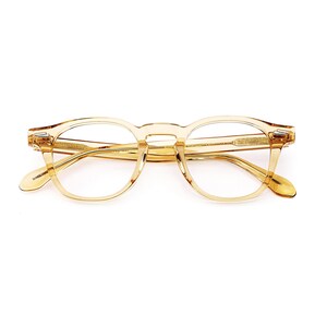 Puede incluir: Un par de gafas de montura redonda transparente, de color &aacute;mbar claro. Las gafas tienen un dise&ntilde;o cl&aacute;sico con un puente de cerradura y detalles sutiles en las patillas. Las lentes son transparentes y la montura es de material transl&uacute;cido.