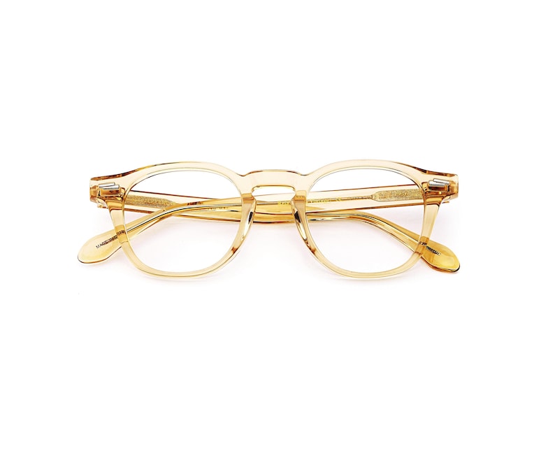 Puede incluir: Un par de gafas de montura redonda transparente, de color &aacute;mbar claro. Las gafas tienen un dise&ntilde;o cl&aacute;sico con un puente de cerradura y detalles sutiles en las patillas. Las lentes son transparentes y la montura es de material transl&uacute;cido.