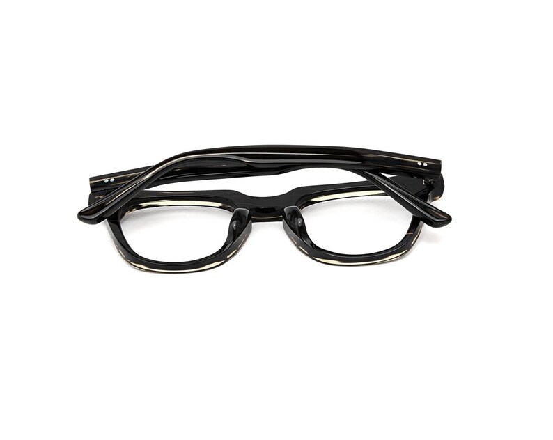 May include: A pair of black eyeglasses with a thick frame and folded temples. The frames feature a glossy finish with a subtle translucent light-colored accent along the bottom edge of the lenses, showcasing a classic rectangular shape.