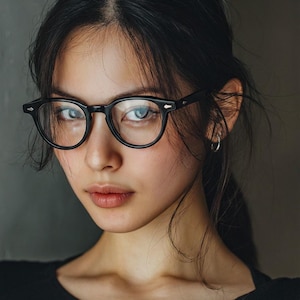 May include: A portrait of a person wearing black, round-framed eyeglasses. The person has dark hair and is looking directly at the camera. The background is a solid, neutral color.