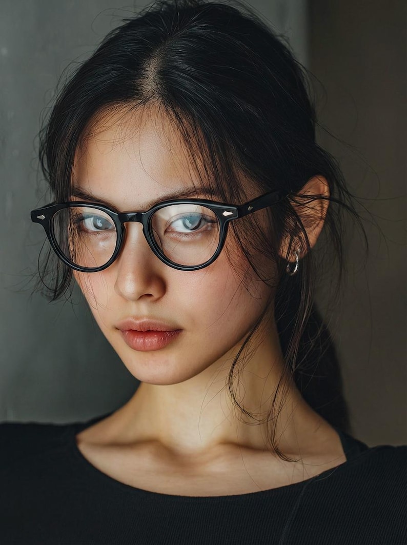 May include: A portrait of a person wearing black, round-framed eyeglasses. The person has dark hair and is looking directly at the camera. The background is a solid, neutral color.