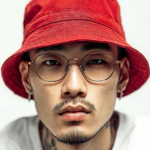 May include: A person wearing a red bucket hat and round eyeglasses. The person has a mustache and goatee. The person is wearing a white shirt. The background is white.