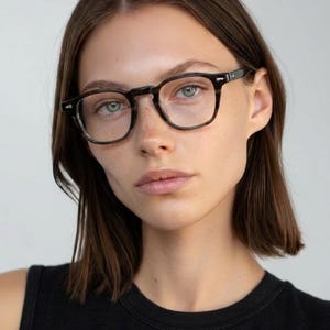 May include: Dark-framed eyeglasses with a square shape. The frames are a dark, mottled color. The model is wearing a black tank top. The background is a plain, light color.