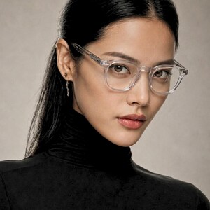 K&ouml;nnte beinhalten: Klare Brille mit quadratischem Rahmen. Die Brille hat einen transparenten Rahmen und wird von einer Person mit dunklem Haar und einem schwarzen Rollkragenpullover getragen. Der Hintergrund ist neutral gef&auml;rbt.