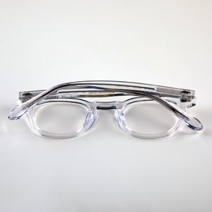 K&ouml;nnte beinhalten: Klare, transparente Brille mit rundem Rahmen. Die Brille ist zusammengeklappt und liegt auf einer wei&szlig;en Oberfl&auml;che. Die B&uuml;gel und der Rahmen sind hell und durchscheinend. Die Gl&auml;ser sind oval geformt.