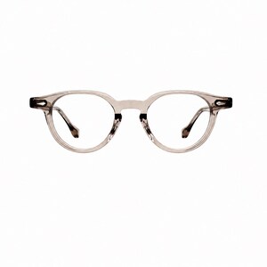 May include: Clear, round-framed eyeglasses with a light gray tint. The frames have a subtle, translucent appearance, and the temples are a darker shade of gray. The lenses are clear, and the overall design is minimalist and modern.