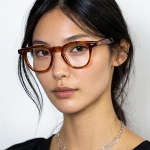 May include: A person wearing brown, square-framed eyeglasses. The glasses have a tortoiseshell pattern on the frames and temples. The person is wearing a black top and a silver necklace.