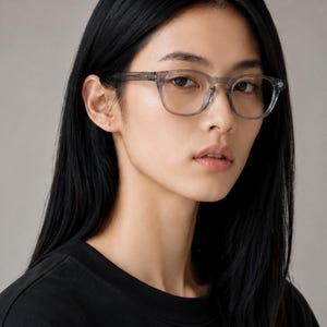 May include: A person wearing clear-framed eyeglasses with a square shape. The glasses have a subtle, translucent gray color. The person is wearing a black crew neck shirt. The background is a solid, neutral gray.