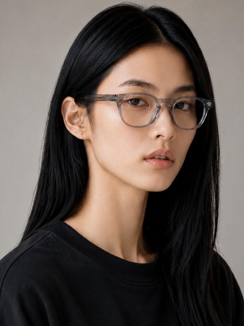 May include: A person wearing clear-framed eyeglasses with a square shape. The glasses have a subtle, translucent gray color. The person is wearing a black crew neck shirt. The background is a solid, neutral gray.