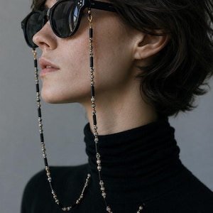 K&ouml;nnte beinhalten: Eine schwarze Sonnenbrille mit einer dekorativen Kette aus abwechselnden schwarzen zylindrischen Perlen, kleinen goldfarbenen runden Zwischenelementen und gesprenkelten wei&szlig;-schwarzen Dalmatiner-Jaspis-Perlen an den Brillenb&uuml;geln.