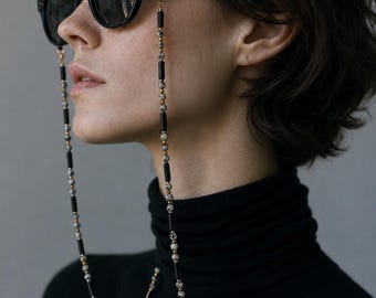 May include: A pair of black sunglasses with a decorative chain featuring alternating black cylindrical beads, small gold-toned spherical spacers, and speckled white and black dalmatian jasper stone beads attached to the frame temples.