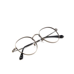 Puede incluir: Gafas redondas con una montura met&aacute;lica fina de color gris plomo y lentes transparentes. El dise&ntilde;o incluye un puente, almohadillas nasales ajustables y varillas delgadas que terminan en puntas de pl&aacute;stico negro liso para mayor comodidad.
