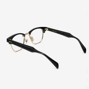 May include: Black and gold browline glasses featuring a semi-rimless frame design with clear lenses. The temples are black with an intricate engraved pattern along the sides. The inner temple is printed with the text 48-20-145 Col.1.