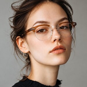 May include: A close-up portrait of a person wearing clear-framed eyeglasses with brown upper rims. The person has brown hair and is wearing a black top. The background is a solid gray.