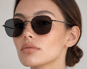 May include: A pair of sunglasses featuring a thin, dark metal frame with a double bridge design and dark, solid-coloured lenses. The eyewear has slender temples and adjustable nose pads, providing a sleek and functional look for eye protection.