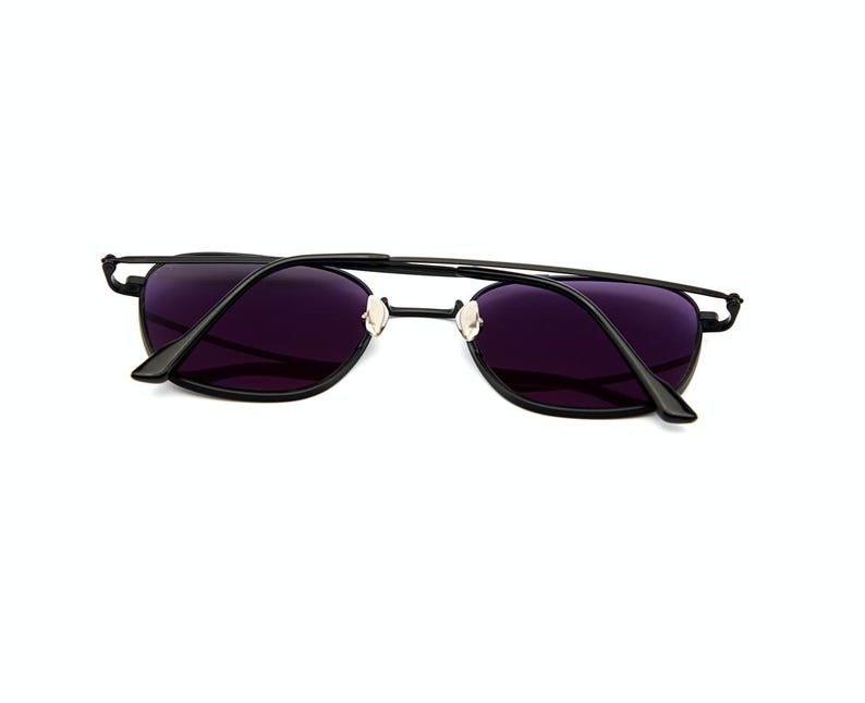 May include: A pair of sunglasses with a thin black metal frame and dark purple tinted lenses. The glasses feature a straight top bar connecting the two rims, adjustable nose pads, and black temple arms that are folded inward toward the center.