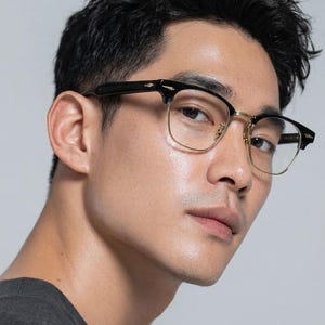 May include: Spectacles featuring a black browline frame with gold-tone metal rims and bridge. The temples are black with a small gold-tone arrow-shaped accent at the hinge. The lenses are clear and the overall design is a classic semi-rimless style.