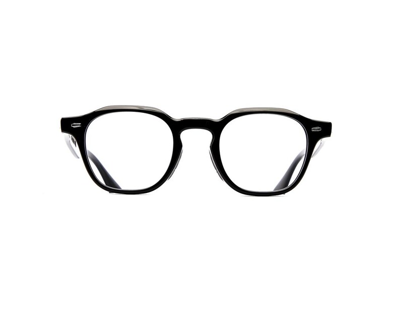May include: Black-framed eyeglasses featuring a rounded, slightly angular shape with a thick bridge and clear lenses. The frames have a glossy finish and include small silver-toned metal accents on the outer corners of the front rims.