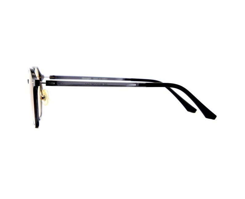May include: A pair of eyeglasses with a black frame and thin temples viewed from the side. The lenses have a light amber tint, and the frame features gold-toned nose pads. The side of the temple includes small printed text detailing the model.