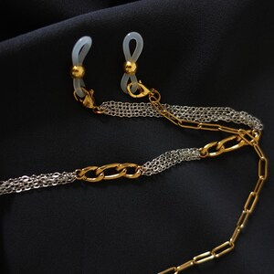May include: A two-tone eyeglass chain featuring alternating sections of gold-colored oval links and silver-colored fine multi-strand chains. The ends include gold-colored beads and translucent silicone loops for attaching to eyewear frames.