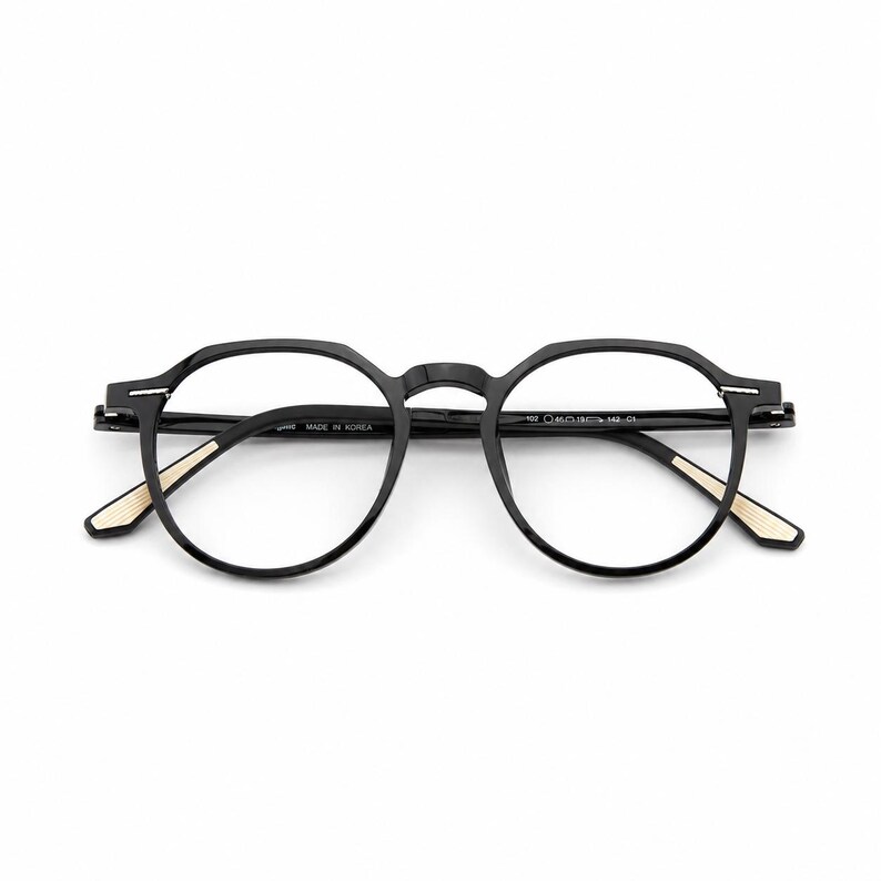 May include: Black eyeglasses with a round frame design. The glasses have a matte finish and feature a subtle gold accent on the temples. The text "MADE IN KOREA" is visible on the frame.