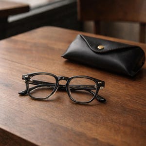 May include: Black-framed eyeglasses with clear lenses rest on a wooden table next to a black leather glasses case. The glasses have a classic, rectangular shape. The case is closed with a gold-colored snap.