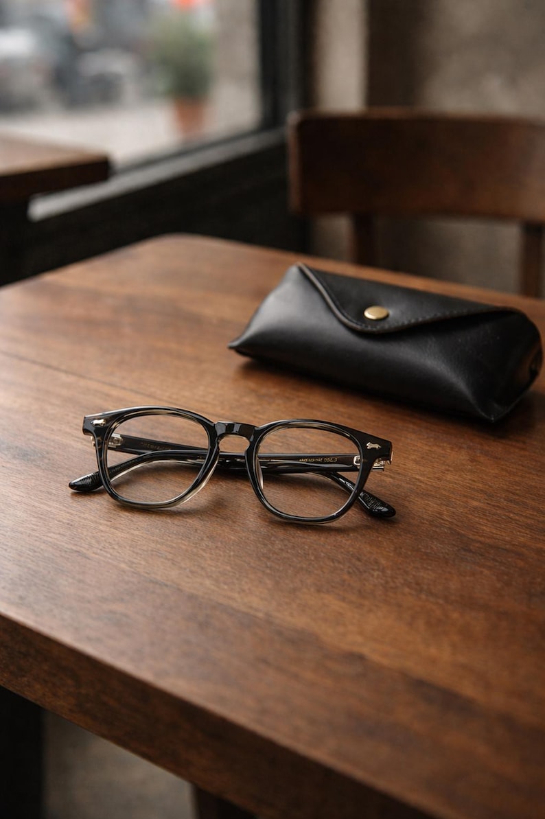 May include: Black-framed eyeglasses with clear lenses rest on a wooden table next to a black leather glasses case. The glasses have a classic, rectangular shape. The case is closed with a gold-colored snap.
