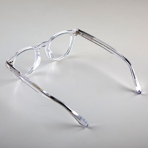 K&ouml;nnte beinhalten: Transparente Brille mit rundem Rahmen. Die Brille besteht aus klarem Material und bietet eine minimalistische &Auml;sthetik. Die B&uuml;gel sind schlank und reichen bis zu den Ohrenteilen. Sie liegt auf einem hellgrauen Hintergrund.