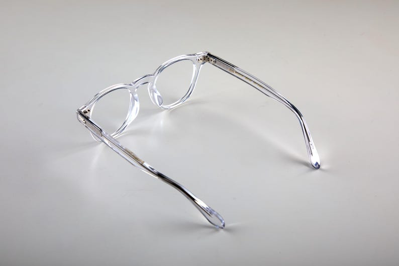 K&ouml;nnte beinhalten: Transparente Brille mit rundem Rahmen. Die Brille besteht aus klarem Material und bietet eine minimalistische &Auml;sthetik. Die B&uuml;gel sind schlank und reichen bis zu den Ohrenteilen. Sie liegt auf einem hellgrauen Hintergrund.