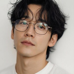 May include: A pair of eyeglasses with thin, metallic, rectangular frames featuring rounded corners. The glasses have clear lenses and are positioned on a face with dark, wavy hair and a white shirt, highlighting the frame's simple design.