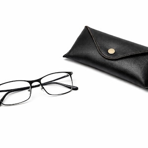 Puede incluir: Gafas rectangulares negras con un estuche de cuero negro a juego. Las gafas tienen una montura fina de metal y lentes transparentes. El estuche se cierra con un broche dorado.