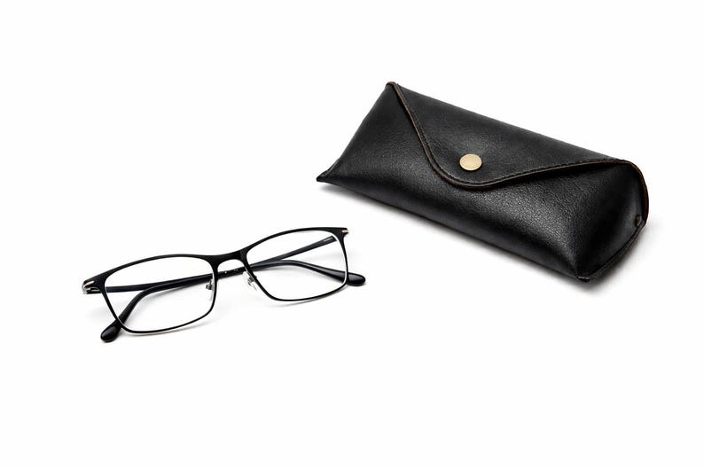 Puede incluir: Gafas rectangulares negras con un estuche de cuero negro a juego. Las gafas tienen una montura fina de metal y lentes transparentes. El estuche se cierra con un broche dorado.